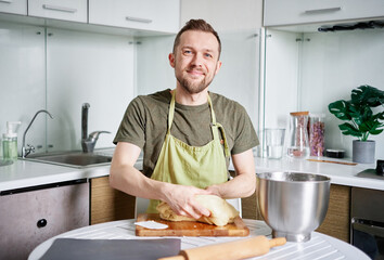 Fototapeta na wymiar Portrait of bearded male chef baker in green apron holding dough on a cutting board with metal bowl. Making pastry or bread at home kitchen. High quality image