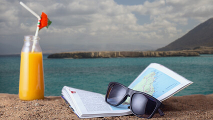 sunglasses with a guide book and a bottle of orange juice on a sandy beach of a warm sea