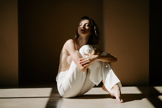 Front view of pensive young girl with naked shoulders and bust, dressed in classic pants, bending legs and holding hands on knees, sitting on floor in studio while sun shining on her face