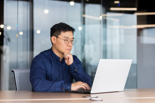 Serious and thoughtful businessman working inside office sitting at table using laptop at work, mature asian boss in shirt thinking and typing on keyboard.