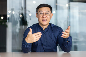 Video call online meeting asian businessman looking web camera and having fun talking and gesturing, man working inside office sharing plans with partners and colleagues, boss in shirt and glasses.
