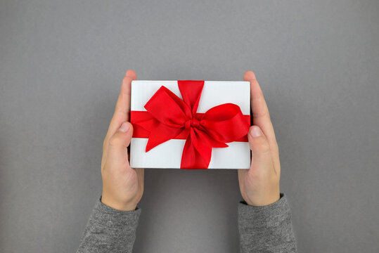 POV Photo, Hands Holding A White Gift Box With A Red Satin Ribbon Bow On An Isolated Gray