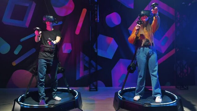 Two joyful, interested young people playing on VR treadmill with helmets and controllers. Slow motion