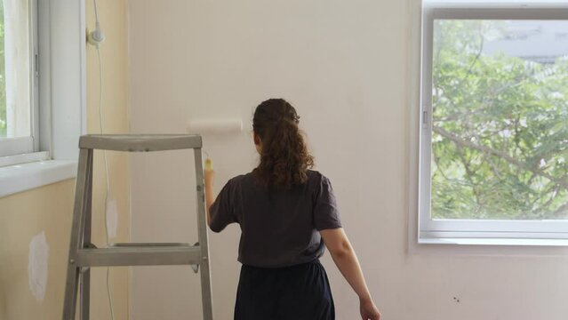 Female is painting a white wall with paint roller at her new apartment or home. Remodeling house or office repainting and doing repair work before moving in or out of place. Ladder next to her