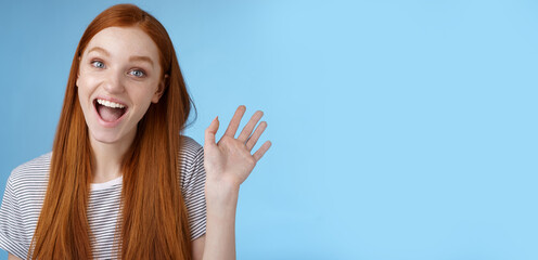 Hello wanna be friends. Enthusiastic cute redhead female newbie getting know coworkers smiling happy waving raised hand hi greeting gesture welcoming, say bye standing blue background - obrazy, fototapety, plakaty