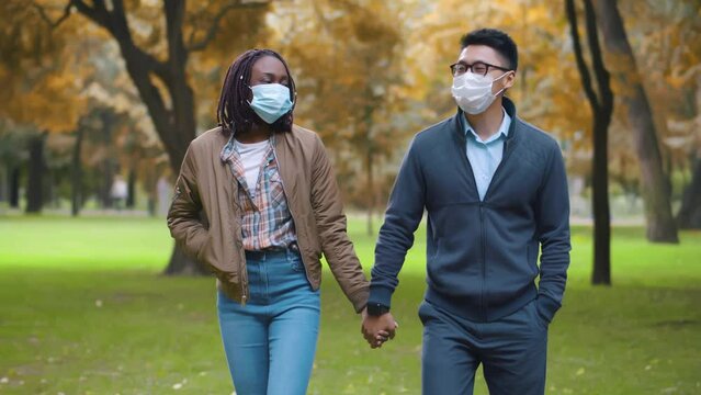 Multiethnic couple in safety mask walk in park. Diverse people in protective mask on date. Realtime