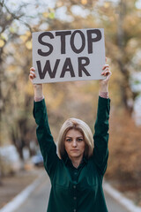 Upset Ukrainian girl protesting war conflict raises banner, placard  with inscription message text Stop War, street background. Crisis, peace, stop aggression, people against Russian invasion concept.