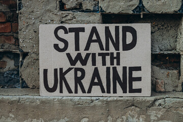Ukrainian protest against war with banner placard  with inscription message text Stand With Ukraine, ruined city background. Crisis, peace, Russian aggression invasion concept. anti-war demonstration.