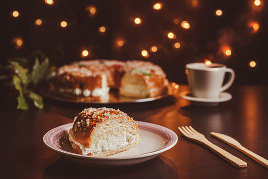 Piece of roscón de reyes on a white and pink plate on a dark table with wooden cutlery, in the background a roscón de reyes and a cup of coffee out of focus and bokeh lights, foreground.
