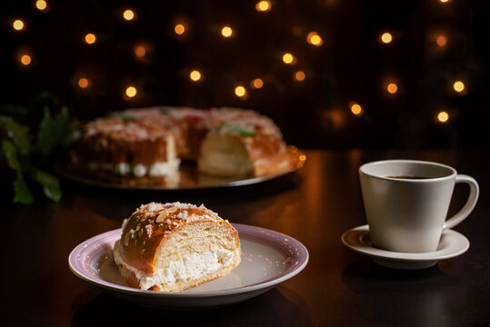 Piece of roscon de reyes next to a cup of coffee, behind a roscon de reyes out of focus and with Christmas lights, foreground.
