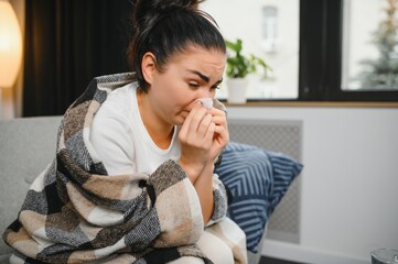 Ill young woman sit on sofa covered with blanket, freezing, blowing her runny nose, sneeze in tissue. Female got fever, flu and influenza symptoms cough at home. Sick allergic with respiratory disease