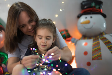 Fototapeta na wymiar Cute little girl interacting interestedly with the Christmas lights, sitting on her mother's lap who teaches her
