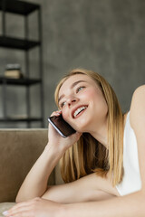 Fototapeta premium Cheerful blonde woman talking on smartphone while lying on couch at home.