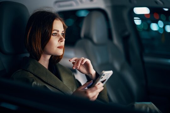  Stylish, Luxurious Woman In A Leather Coat Sitting In A Black Car At Night On The Passenger Seat, Looking Thoughtfully To The Side, Holding Her Hand Near Her Face, And The Other Holding A Smartphone