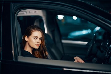 Obraz na płótnie Canvas a stylish, luxurious woman sits in a black car at night in the passenger seat, and looks pleasantly into the camera. Close horizontal photo