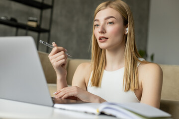 Young coach in wireless earphone holding pen during video chat on laptop near blurred notebook at home.