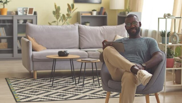 Portrait of African American freelancer using digital tablet while sitting in armchair in living room, working from home