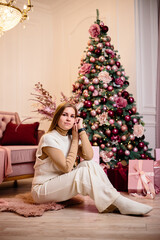 Obraz na płótnie Canvas A stylish happy young woman in a fashionable white knitted suit is sitting on a soft rug in a cozy room near a Christmas tree. Fashionable girl joyful model rests in the Christmas studio