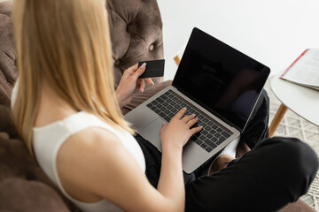 Blurred young woman holding credit card and using laptop with blank screen on couch at home.