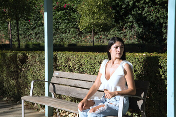 Naklejka premium South American woman, young and beautiful, brunette, with white shirt and jeans, sitting on a wooden bench, alone, calm and relaxed. Concept beauty, fashion, solitude, tranquility, relaxation.