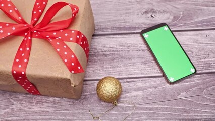Golden christmas ball rolling towards smart phone mockup with chroma key, blank green screen on wooden table with gift boxes. Layout for advertising goods and services in a mobile application. - Powered by Adobe