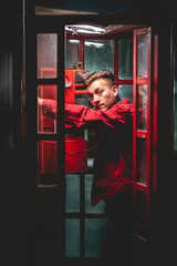 Fototapeta na wymiar Portrait of young and attractive model guy with red jacket smoking a cigarette in a red phone booth in the city in the night