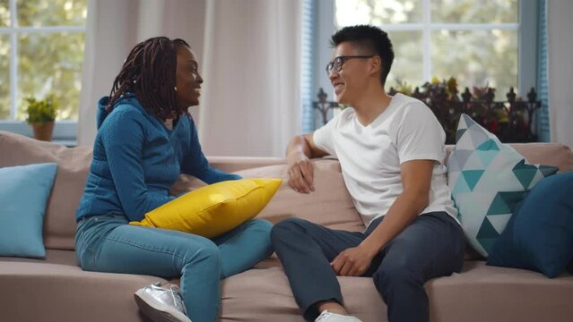Multiethnic couple talk on couch. Diverse friends chat and relax on sofa. Realtime