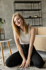 cheerful blonde woman in white tank top and black jeans looking at camera on floor at home.