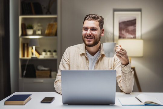 Happy young man holding cup of hot everage and smiling to camera, sitting at desk with laptop. Remote developer finishing project