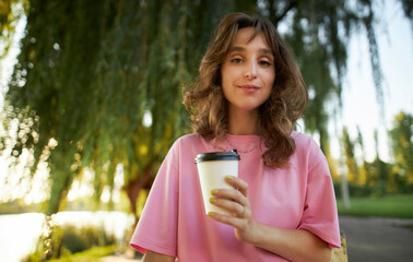 Photo of cute charming young lady in pink t-shirt, beaming smile, holding paper coffee cup, park outdoors. Cheerful young girl in a pink t-shirt, walking in the park, holding a cup of takeaway coffee