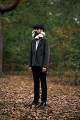 Fototapeta na wymiar The surreal theme - a bearded man with a twisted moustache, in a blazer and hat with a bouquet of white flowers around his neck, in forest.