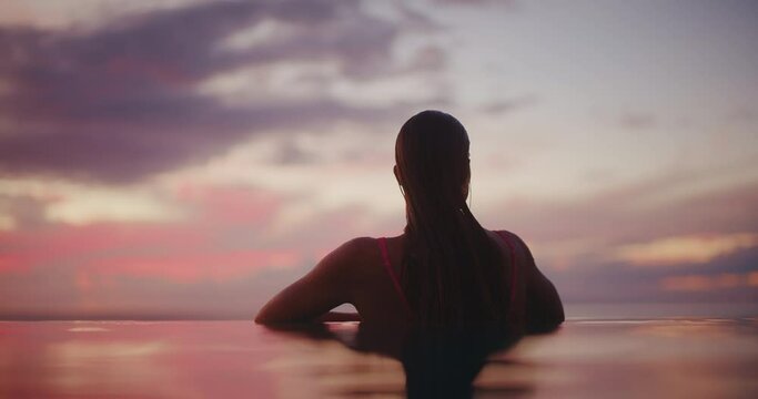 Woman relaxing in luxury infinity pool, looking out over the ocean at sunset, tropical resort spa vacation