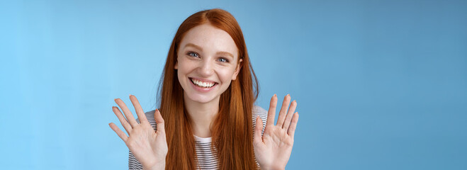 Naklejka na ściany i meble Charming redhead elder sister say goodbye sibling friends smiling cheerful waving raised palms show ten fingers grinning joyfully look carefree relaxed, talking casually blue background