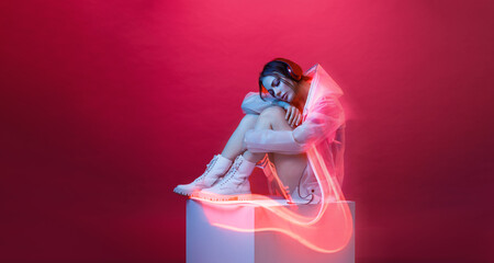 Young woman in modern headphones listens to music. Woman in futuristic costume. Augmented reality game, future technology, AI concept. VR. Neon red light. Viva magenta color of the year 2023. 