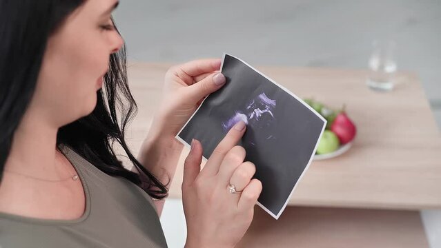 young mother is expecting baby, adorable pregnant woman is looking at photo of an ultrasound in her hands, close-up plan
