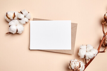 Holiday greeting card mockup with dried branch of cotton flowers and envelop on beige background, top view, flat lay. White wedding invitation card mock up