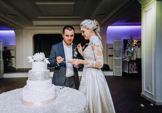 Stylish groom and beautiful blonde model bride eat delicious sweet cake while feeding each other. Wedding photography of newlyweds, food.
