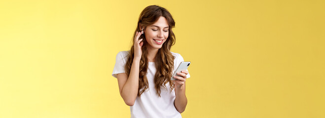 Tender feminine curly-haired female white t-shirt touch wireless earbud put earphone ear smiling pleased look smartphone screen pick song wanna listen music search right track yellow background