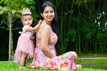 portrait of latina mother sitting on grass with her beautiful brunette baby girl standing touching her back, learning to walk