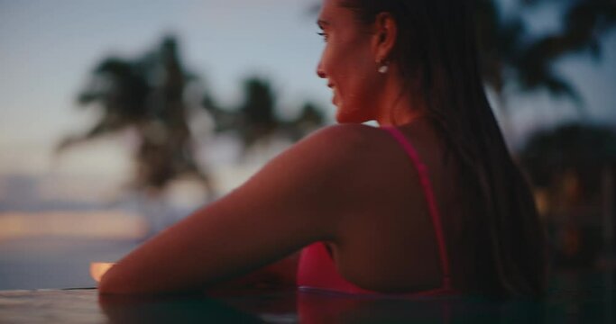 Close up of beautiful young woman relaxing in the pool at tropical resort on vacation, looking out over the ocean at sunset