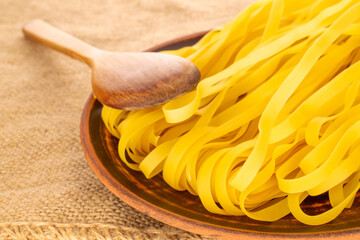 Homemade raw noodles in clay plate with wooden spoon on jute cloth, macro.