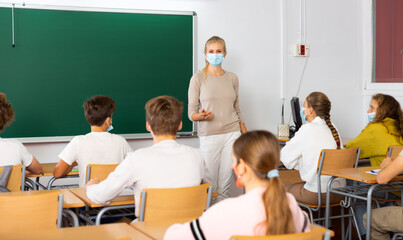Female speaker in protective mask giving lesson for teenage students in classroom