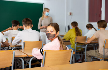Young girl in face mask turned around and looking at camera while sitting at desk during lesson in classroom.
