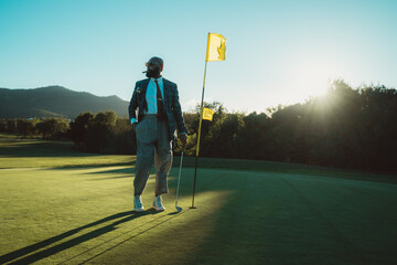 A bald bearded man, with an old money style, wears sunglasses, smokes a cigar, and poses with a golf club in his hand, near a flag-stick on a sunny day, in a lush green golf field with shrubs