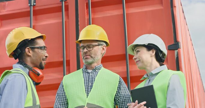 Portrait of three professional hard-working people working on cargo transportation company. Middle-aged boss instructing African man and young woman wearing helmets near red container.