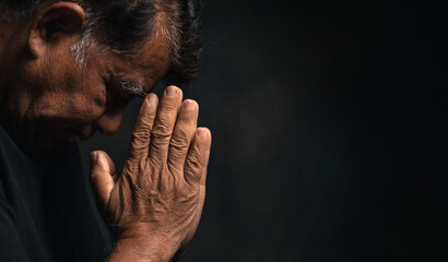 Elderly Asian man bowed his head praying to God on a black background at home. Holding hands in prayer, eyes closed. - obrazy, fototapety, plakaty