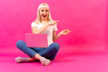 Obraz na płótnie Canvas Blonde caucasian girl with a computer, studio shot on pink background, copy space, pointing