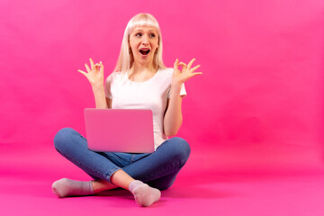 Obraz na płótnie Canvas Blonde caucasian girl with a computer, studio shot on pink background, copy space, ok gesture