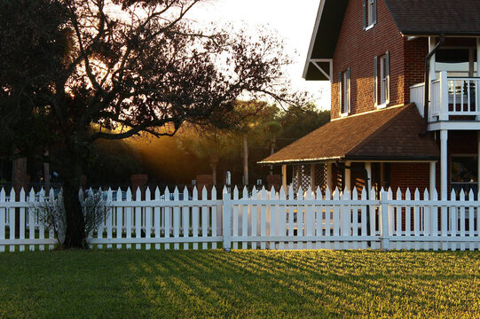 Light shining through the trees at sunset onto a beautiful brick home with a white picket fence. 