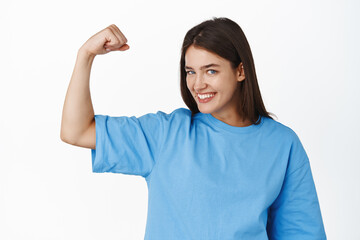 Portrait of smiling girl in t-shirt flexing biceps, showing arm muscle and looking pleased, going workout in gym, standing over white background